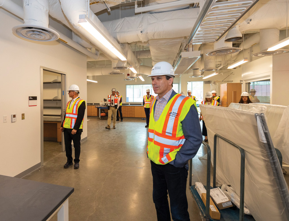 A group of people, including construction workers and university officials, wear hard hats and safety vests while touring a new, bright, and open-plan laboratory or academic facility under construction. The flooring is concrete, and the exposed ceiling has ductwork.