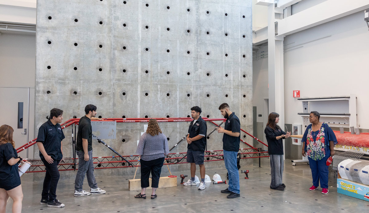 Students and faculty gather in a large concrete laboratory space to examine a massive, tall, concrete reaction wall with a grid of circular holes. Red steel bracing is set up against the wall, and construction materials are scattered on the floor.