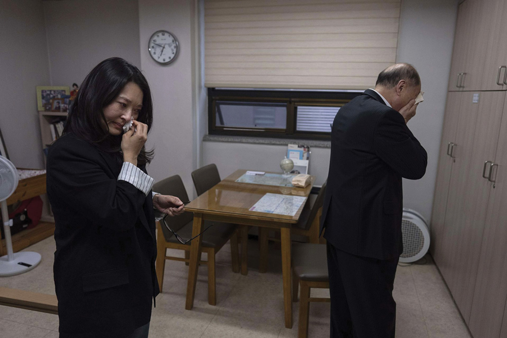 A man and a woman, both dressed in black suits, stand in a room wiping tears from their faces, conveying deep distress or mourning.