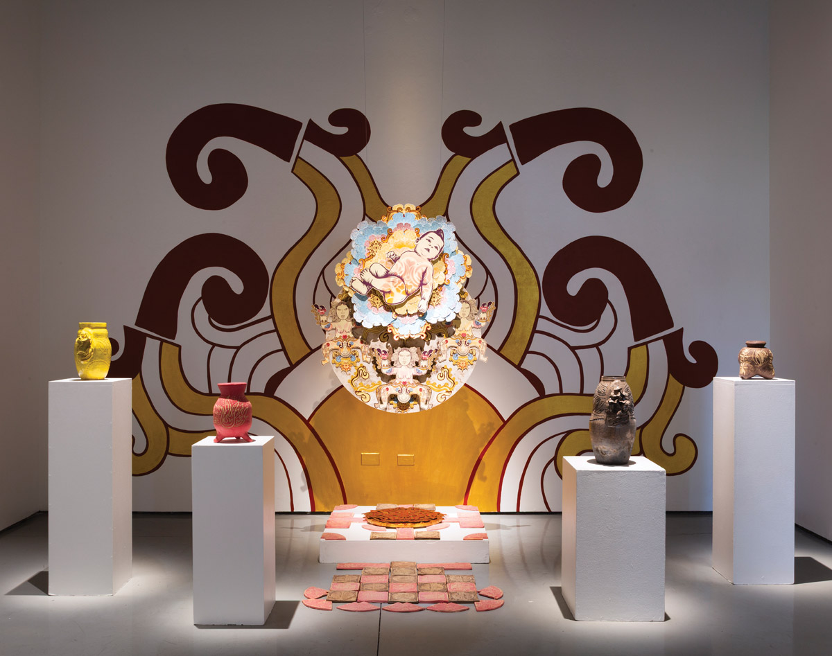 A contemporary art installation featuring a central circular, layered mixed-media piece depicting a figure, possibly religious or mythical, in light colors. This is set against a large wall mural with stylized, swirling designs in deep red-brown and gold-yellow. In the foreground, four ceramic vessels of different colors and textures rest on white pedestals, two on the left and two on the right. Smaller, reddish-pink textured tiles are arranged on the floor around a central lower platform.