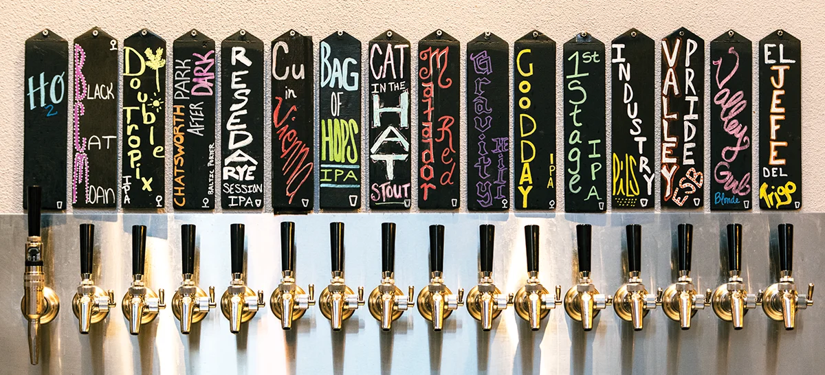 A row of 15 chalkboard tap handles displays the brewery's current beer selections, including 'Matador Red,' 'Reseda Rye,' and 'Valley Girl Blonde,' above a line of brass beer taps.
