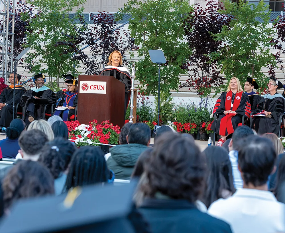 A woman in a commencement gown and hood stands at a podium with the CSUN logo, speaking to a large audience of graduates and spectators.