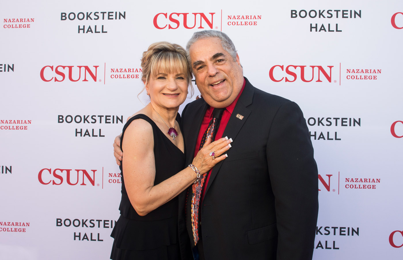 A smiling man and woman embrace on a red carpet in front of a backdrop branded with "CSUN," "Bookstein Hall," and "Nazarian College."