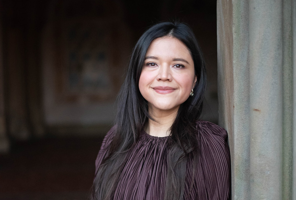Headshot of a smiling woman with long dark hair, wearing a dark pleated top, standing next to a stone column.