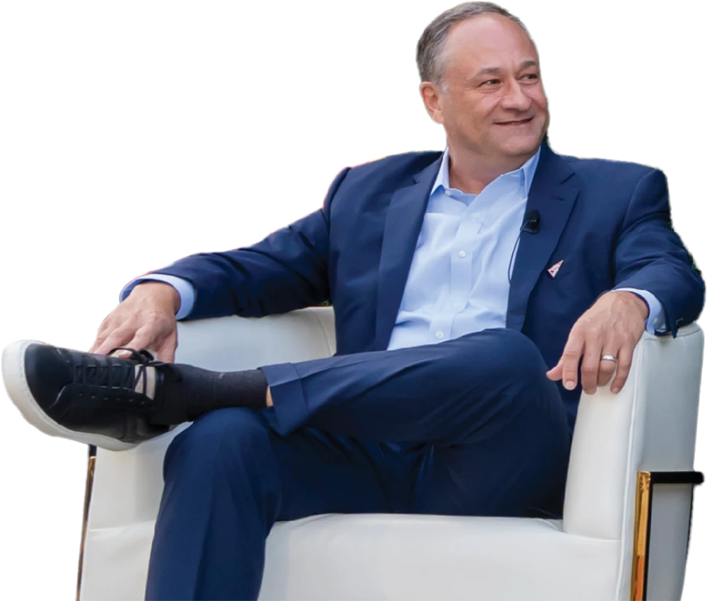 A middle-aged man in a blue suit and light blue shirt is sitting in a white armchair with his legs crossed, smiling and looking off to the side. He is wearing black sneakers with white soles.