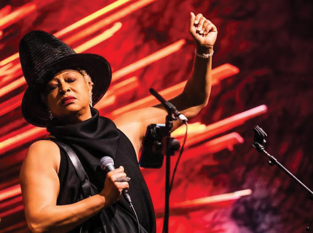A woman wearing a large, structured black hat and black sleeveless outfit is performing on stage. She is holding a microphone in her right hand and has her left arm raised in the air against a dramatic red and orange background.