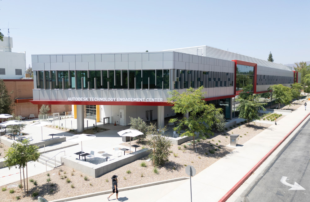 Exterior view of the modern, glass and gray paneled 'Autodesk Technology Engagement Center' building with an outdoor plaza, trees, and patio seating.