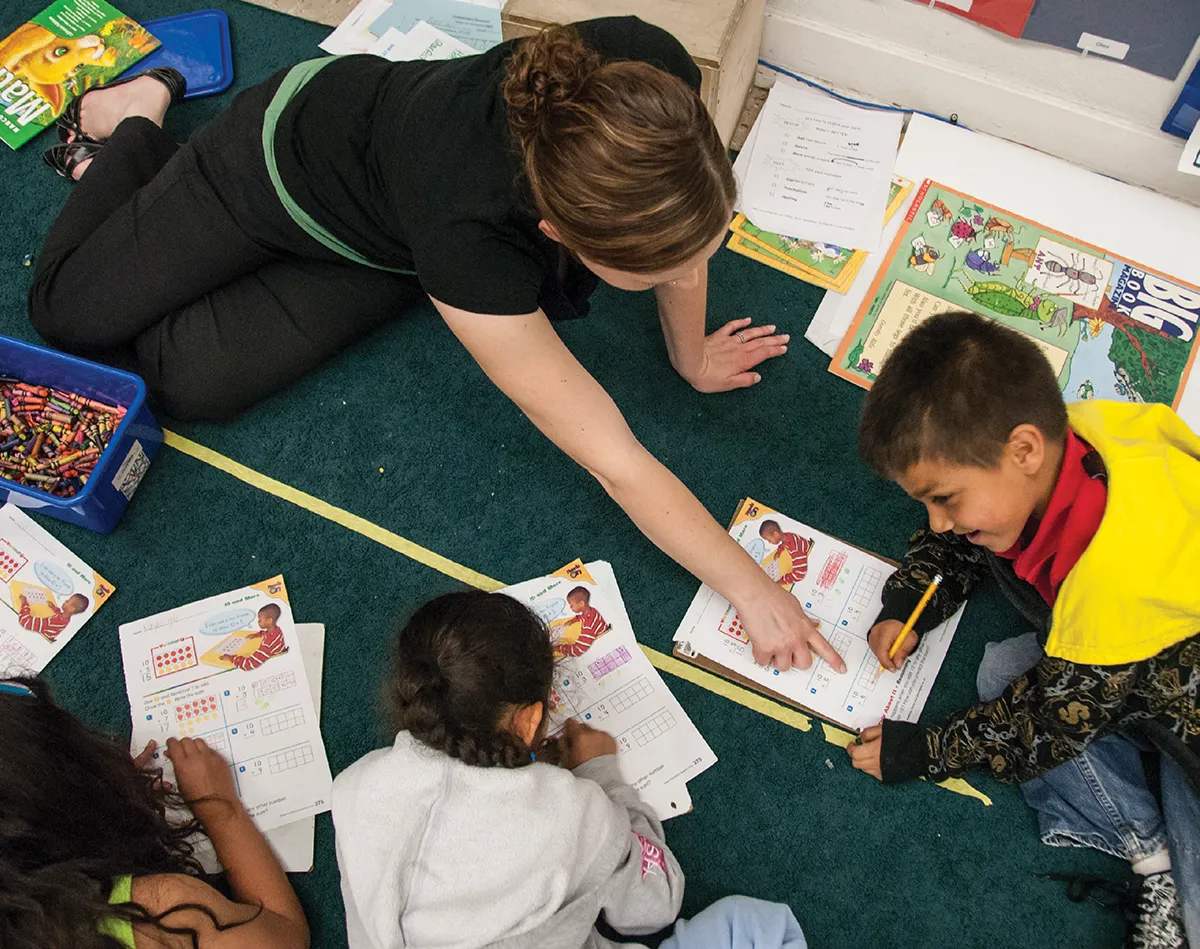 A high-angle view of a female teacher sitting on a green carpet, leaning in to help two young elementary school students with their math worksheets. A box of crayons is visible nearby.