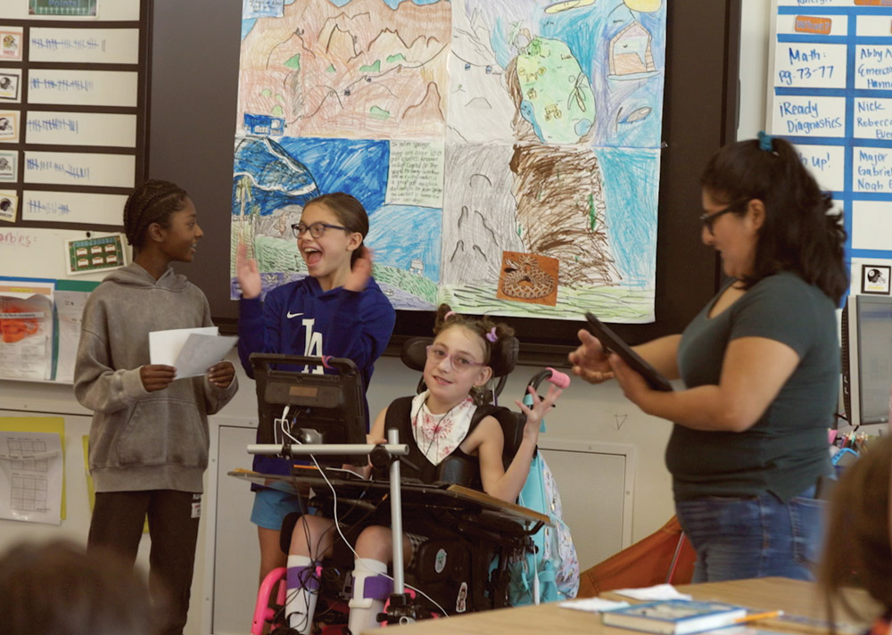 Four people in a classroom presenting a project. A young girl is seated happily in a mobility device, high-fiving another student. Two other students stand nearby, one holding papers and a teacher is holding a tablet, all standing in front of a large, colorful, student-made mural.