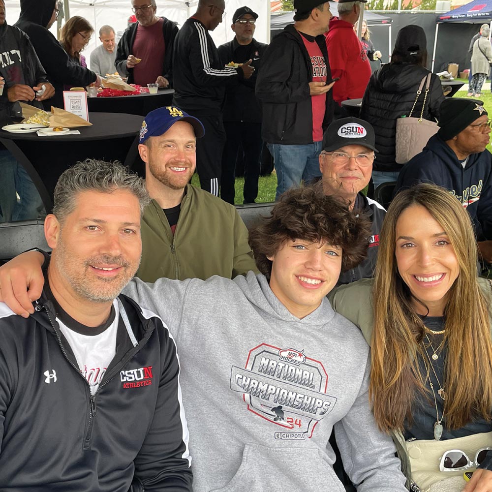 A family of four smiling and posing for a selfie under a tent at an outdoor event. A man on the left is wearing a black and white "CSUN Athletics" zip-up jacket. The teenage boy in the center is wearing a gray hoodie.