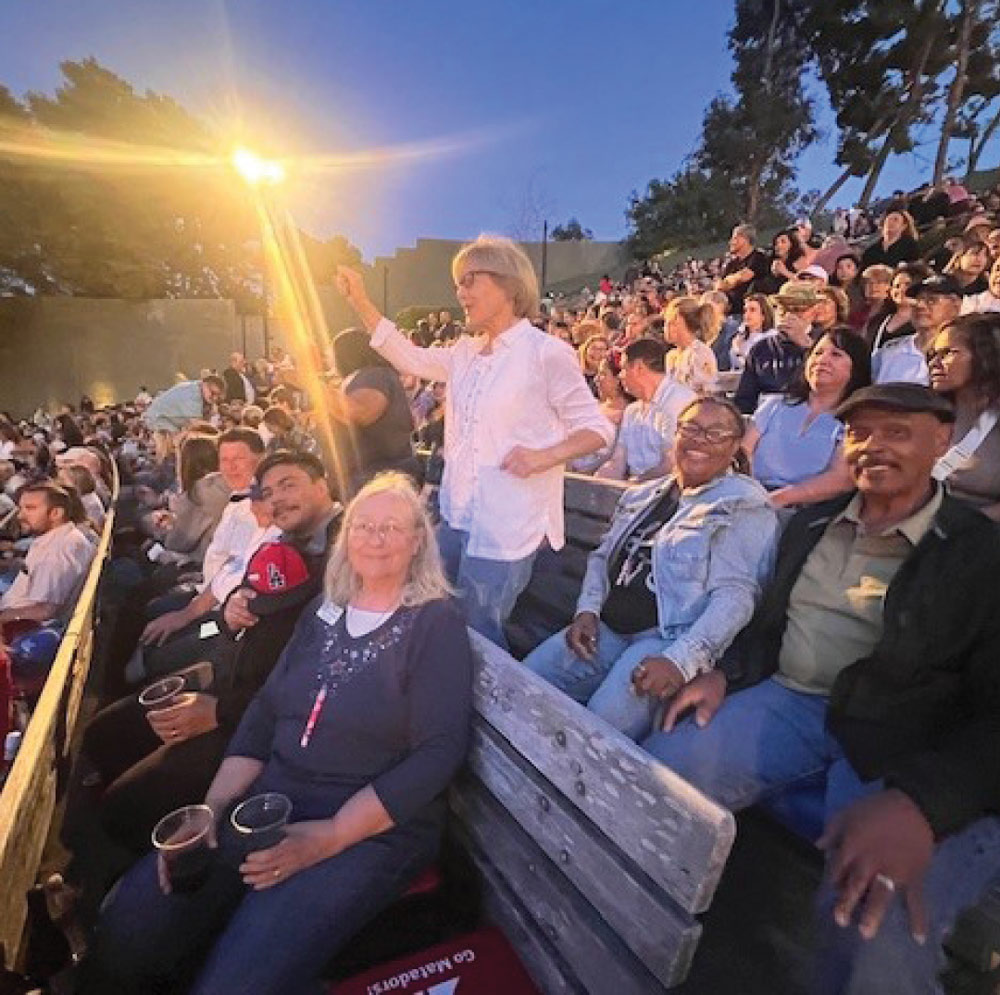 A crowd seated in wooden bleachers at an outdoor event at dusk. A woman in a white shirt stands in the aisle with her hand raised. People in the foreground are smiling and enjoying the event.