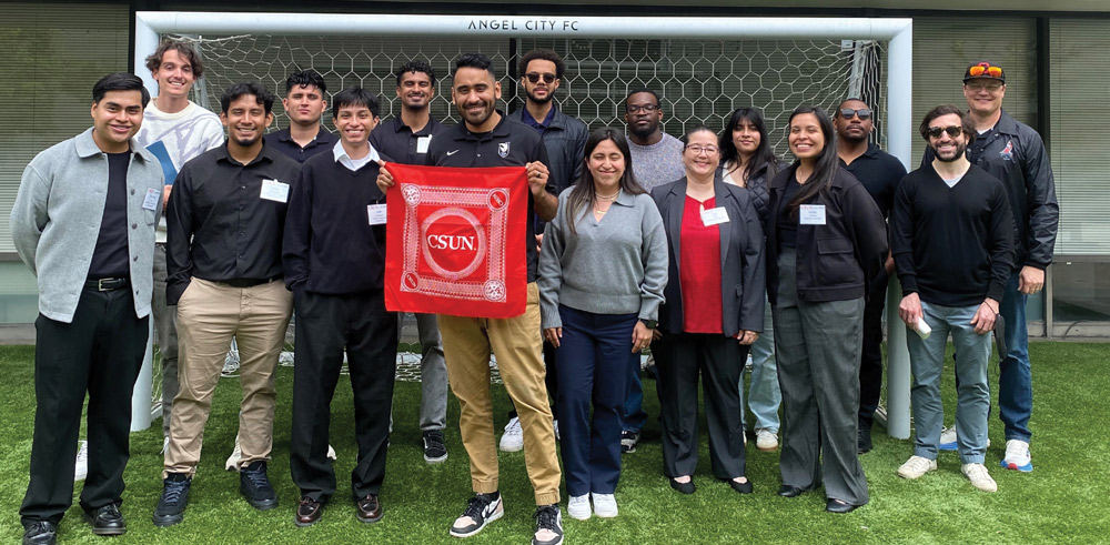 A group of approximately 16 people, a mix of men and women, standing on a grassy field in front of a soccer goal net with an "Angel City FC" banner above. A man in the center holds a red bandana with "CSUN" on it. Most people are wearing business casual or black and gray clothing.