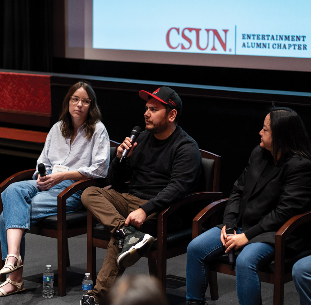 Three people sitting on a panel on a stage. A man in a black shirt and red baseball cap is holding a microphone and speaking, flanked by two women. A screen in the background shows a "CSUN Entertainment Alumni Chapter" logo.