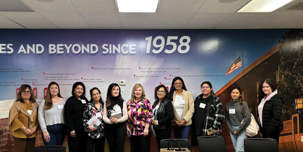 A group of 11 women standing in front of a wall mural detailing the history of the university. The women are dressed in business casual attire and appear to be attending a meeting or event.