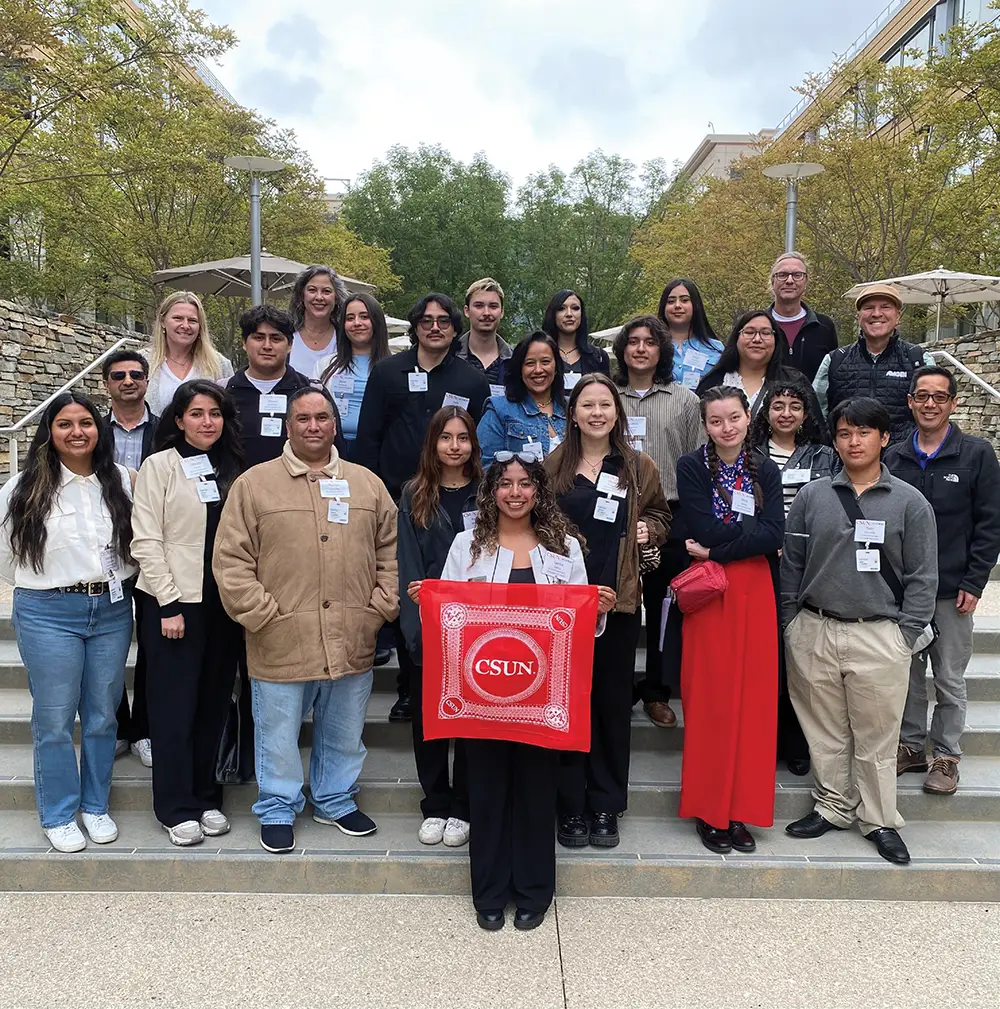A large group of college-aged students and adults standing on wide stone steps outdoors. A student in the center foreground holds a red bandana with "CSUN" on it. They are posing in front of modern campus buildings with trees and bushes.