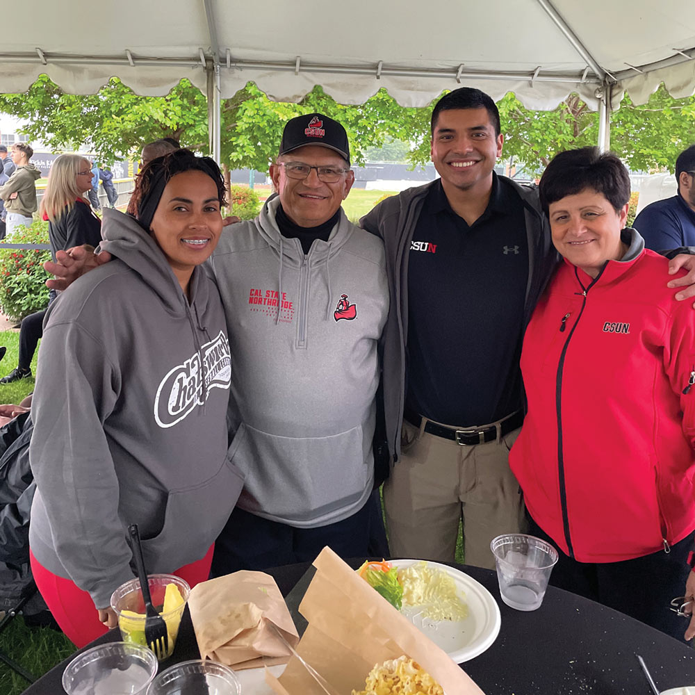 Four people smiling and posing for a photo under a tent at an outdoor event. All four appear to be wearing CSUN-branded apparel, including a man in a gray hoodie and a woman in a red jacket. Food and plastic cups are on the table in the foreground.