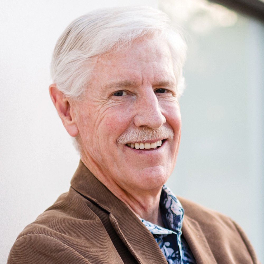 Headshot of a smiling, older white man with white hair and a mustache, wearing a brown corduroy jacket and a patterned shirt. He is leaning against a white wall.
