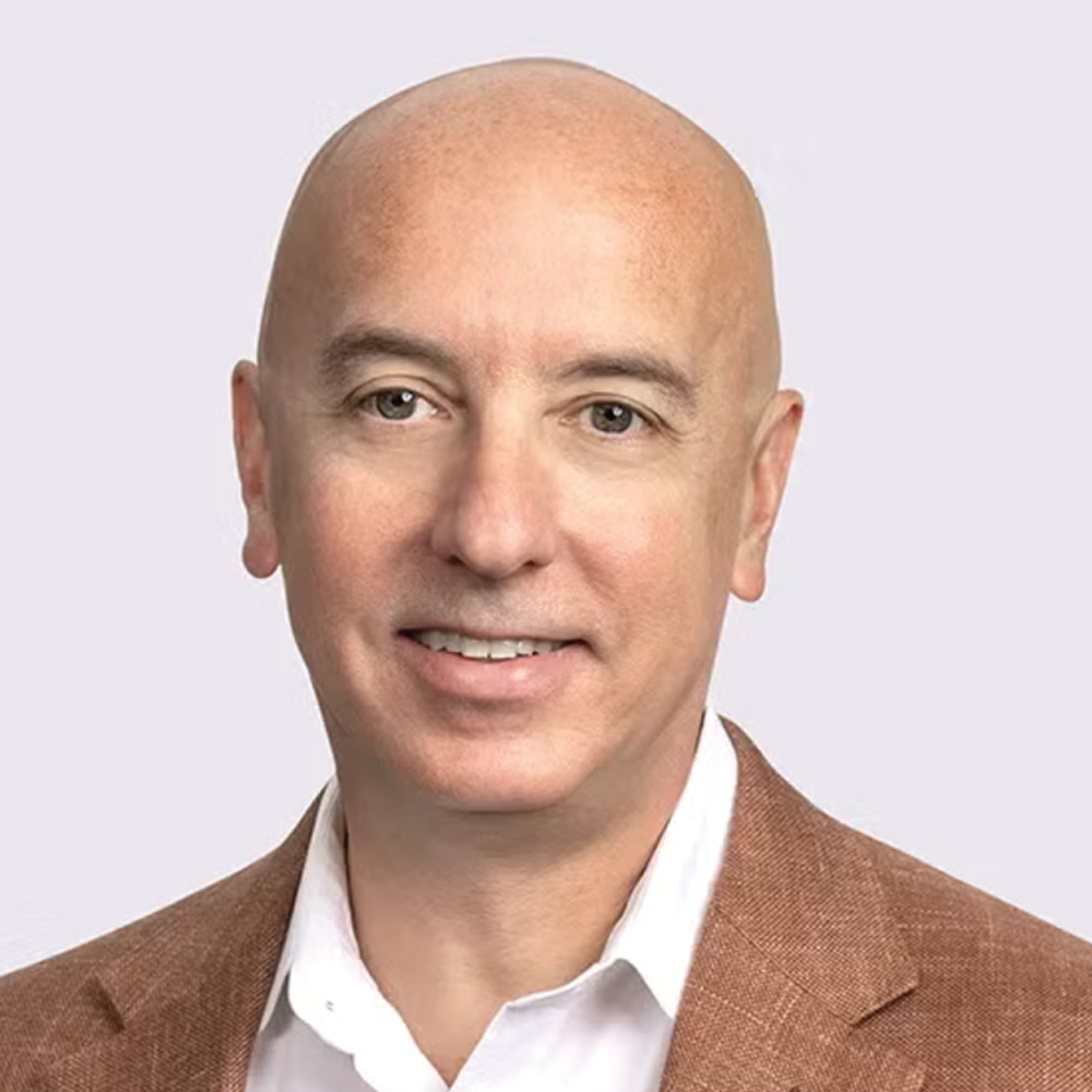 Professional headshot of a smiling, bald white man wearing a light brown or tan blazer over a white collared shirt. The background is a plain light gray.