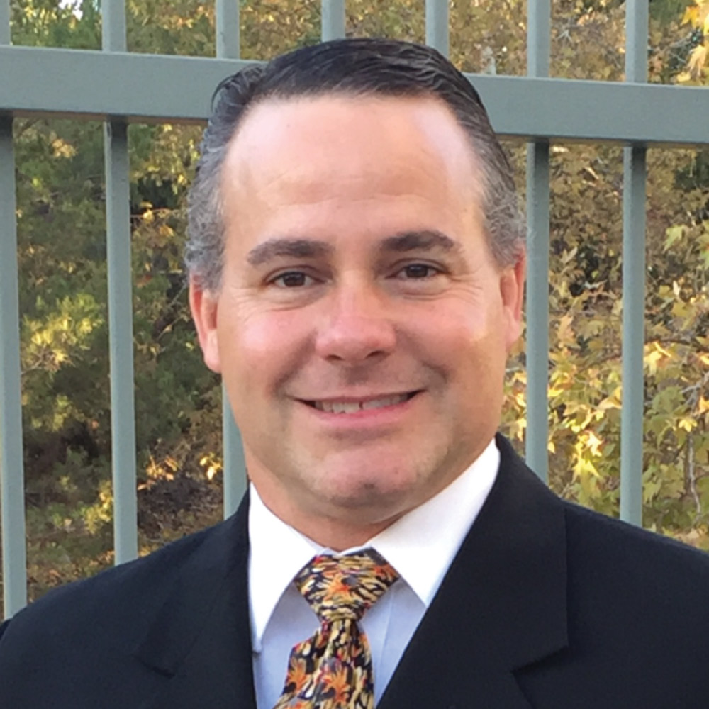 Headshot of a smiling man with dark hair, wearing a black suit jacket and a white shirt with an orange and black patterned tie. He is standing outdoors in front of a metal fence and autumn foliage.