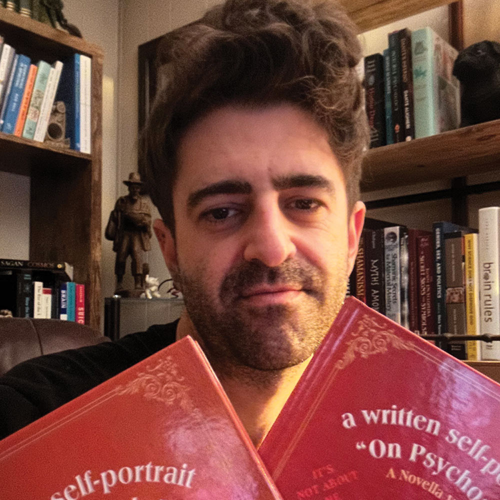 A man with dark, messy hair is holding two red books in front of his face, titled 'a written self-portrait' and 'On Psychology.' He is smiling slightly, with bookshelves visible behind him.