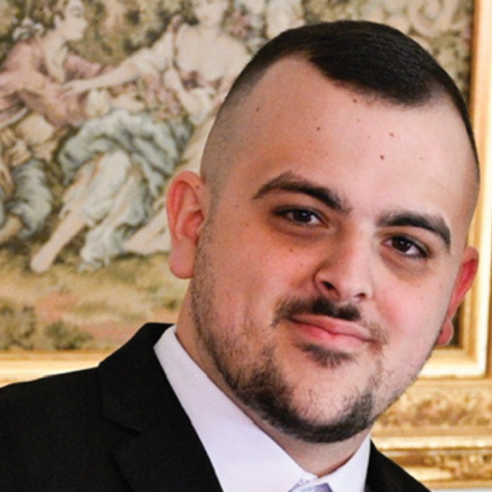 Headshot of a man with a dark beard and a high-fade haircut, wearing a black suit jacket and white shirt, with a neutral-colored painting of people in the background.