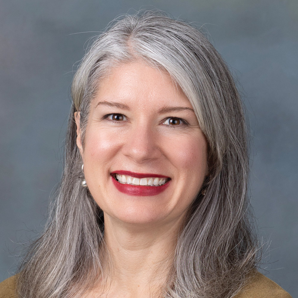 Professional headshot of a smiling woman with long, straight gray hair and bright red lipstick, wearing pearl earrings and a tan top. The background is a muted blue and gray gradient.