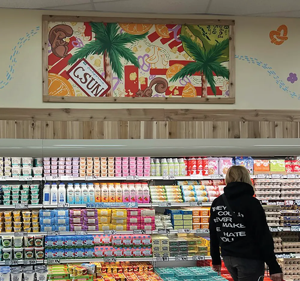 A person stands in a grocery store aisle looking at refrigerated dairy cases, beneath a colorful mural featuring palm trees, squirrels, and the letters CSUN.