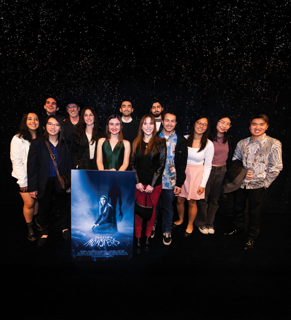 A group of thirteen young people, including filmmakers and a cast, pose together with a movie poster for "Dahlia’s Monsters" at a film premiere.