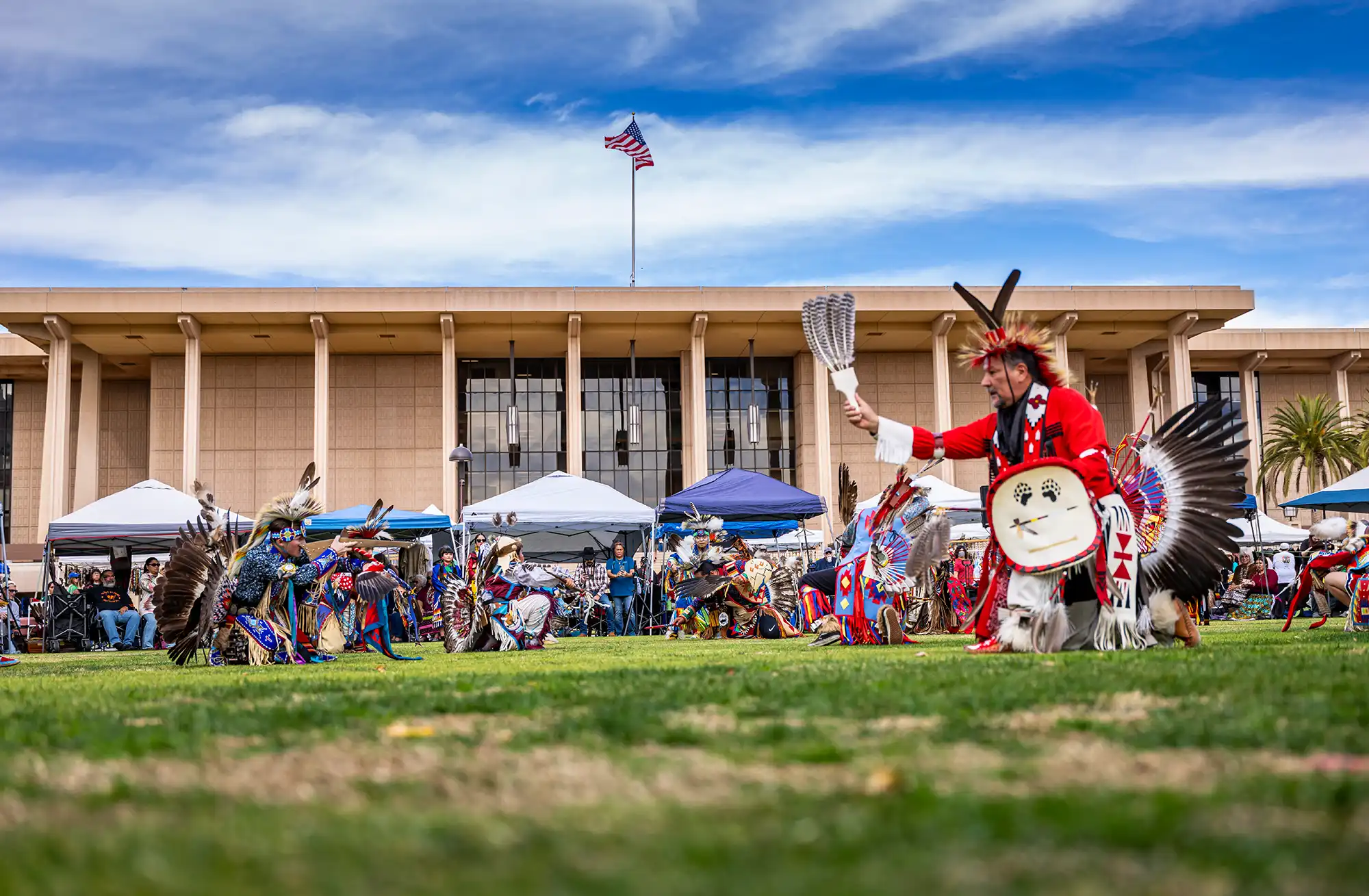 Native American dancers in vibrant traditional regalia perform on a green lawn during a powwow, with an institutional building, vendor tents, and an American flag flying in the background.