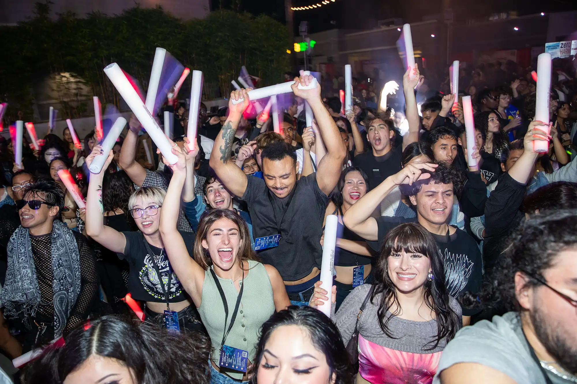 A diverse crowd of young people cheering and holding up white glow sticks or LED foam tubes at a lively outdoor night event. The atmosphere is energetic, with many people smiling and raising their arms.