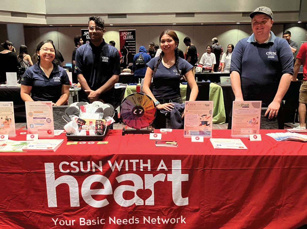 Four students smile behind a red table at a resource fair for "CSUN With A Heart: Your Basic Needs Network," which includes a spinning prize wheel and informational displays.