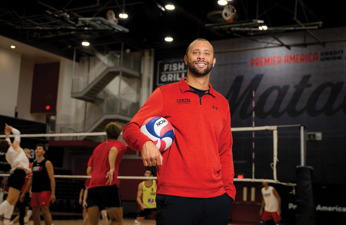 Headshot of a smiling man, presumably a coach, in a red CSUN shirt, holding a volleyball under his right arm. He is standing in a large indoor gymnasium, with volleyball players practicing in the blurred background.