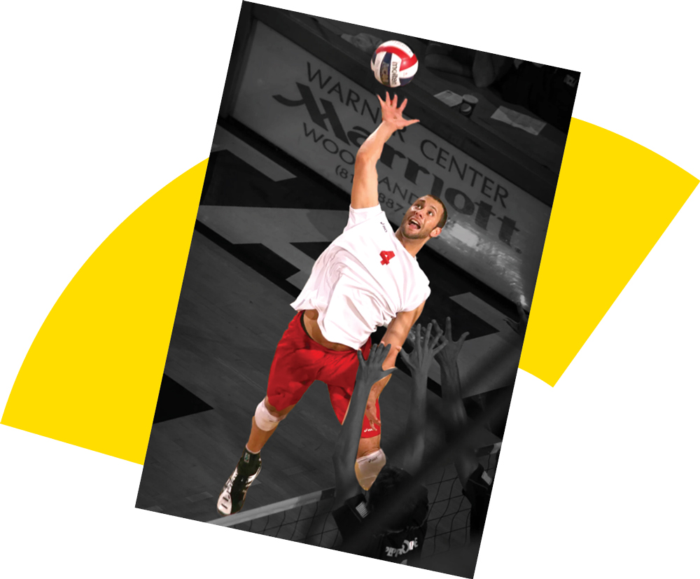 A dynamic, colorized image of a male volleyball player in mid-air, wearing a white jersey and red shorts, spiking the ball over the net. Opposing hands are blocking below him. The background is a black and white blur of the gym.