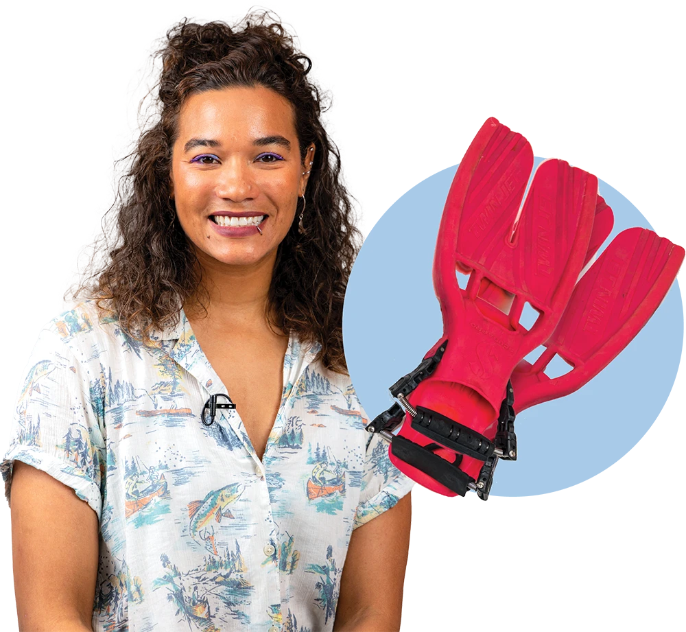 A smiling woman with curly hair wearing a light printed shirt featuring fishing and boating scenes. Next to her is a circular inset image showing a pair of red diving fins.
