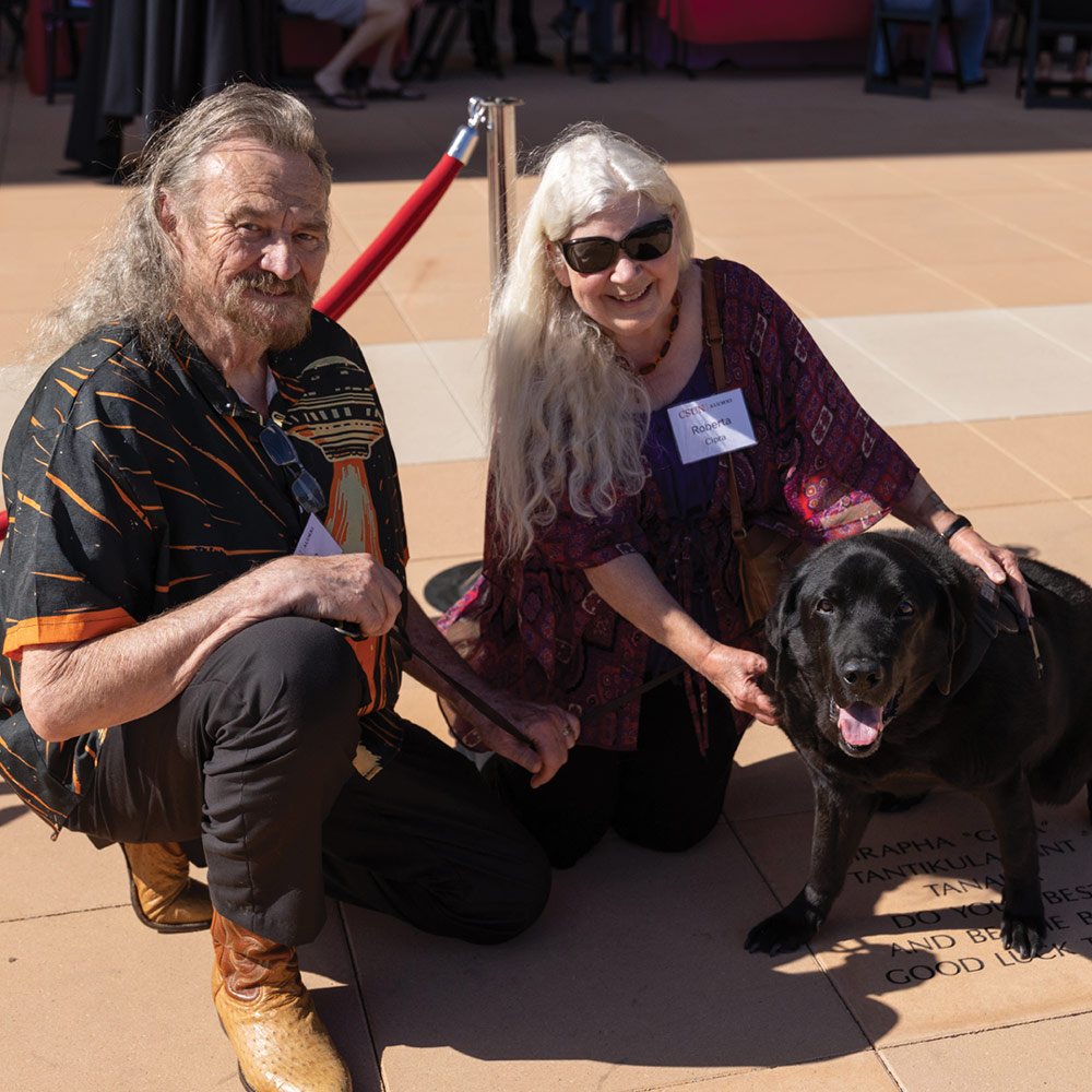 An older couple kneeling on a patio and smiling with their black Labrador retriever.