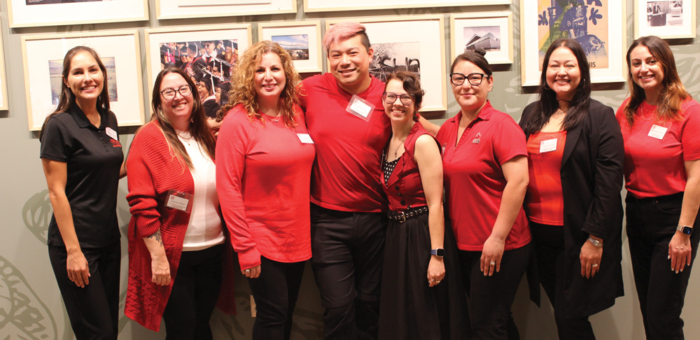 A group of women dressed in red posing together in a gallery space with framed photos on the wall.