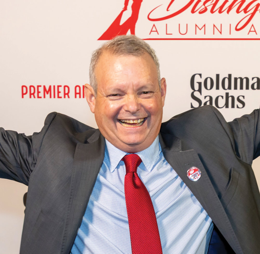 An older man in a grey suit and red tie smiles widely with his arms raised in celebration.
