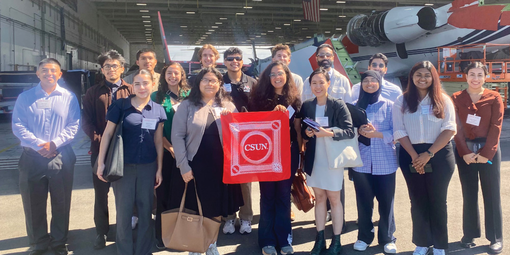 A diverse group of students standing in an aircraft hangar, holding a red CSUN bandana before a plane.