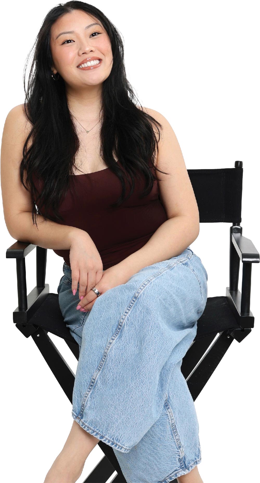 A smiling young woman with long dark hair sits in a black director's chair wearing a brown top and jeans.