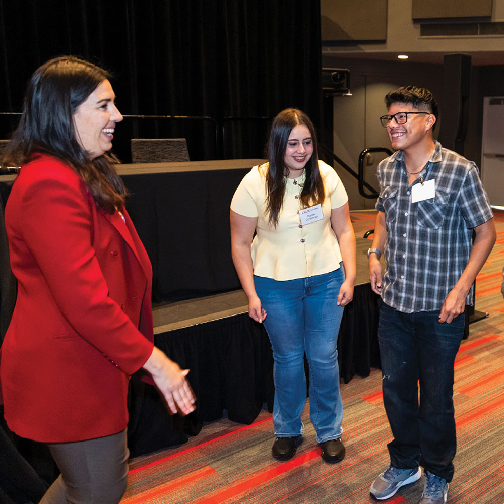 A woman in a red blazer smiles while speaking with two students at an event in an indoor venue.
