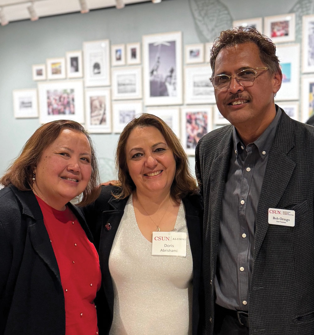 Three people smiling and posing for a group photo in front of a gallery wall covered with framed photographs.