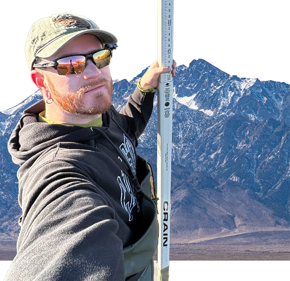 A bearded man wearing a cap, sunglasses, and a hoodie holds a measuring rod in front of a large, snowy mountain range.