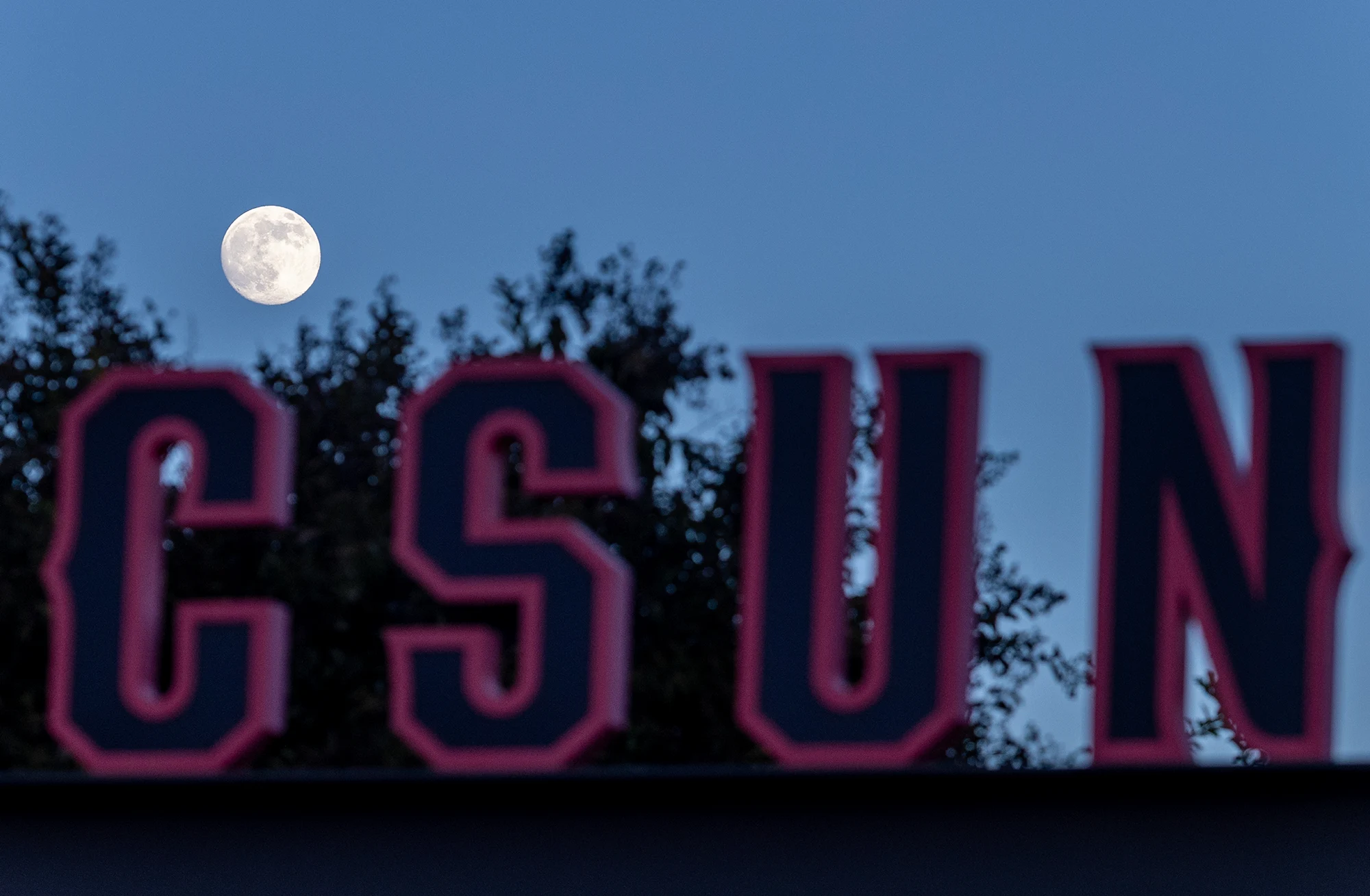 Large red letters spelling "CSUN" stand in the foreground with a bright, full moon glowing in the blue twilight sky behind.