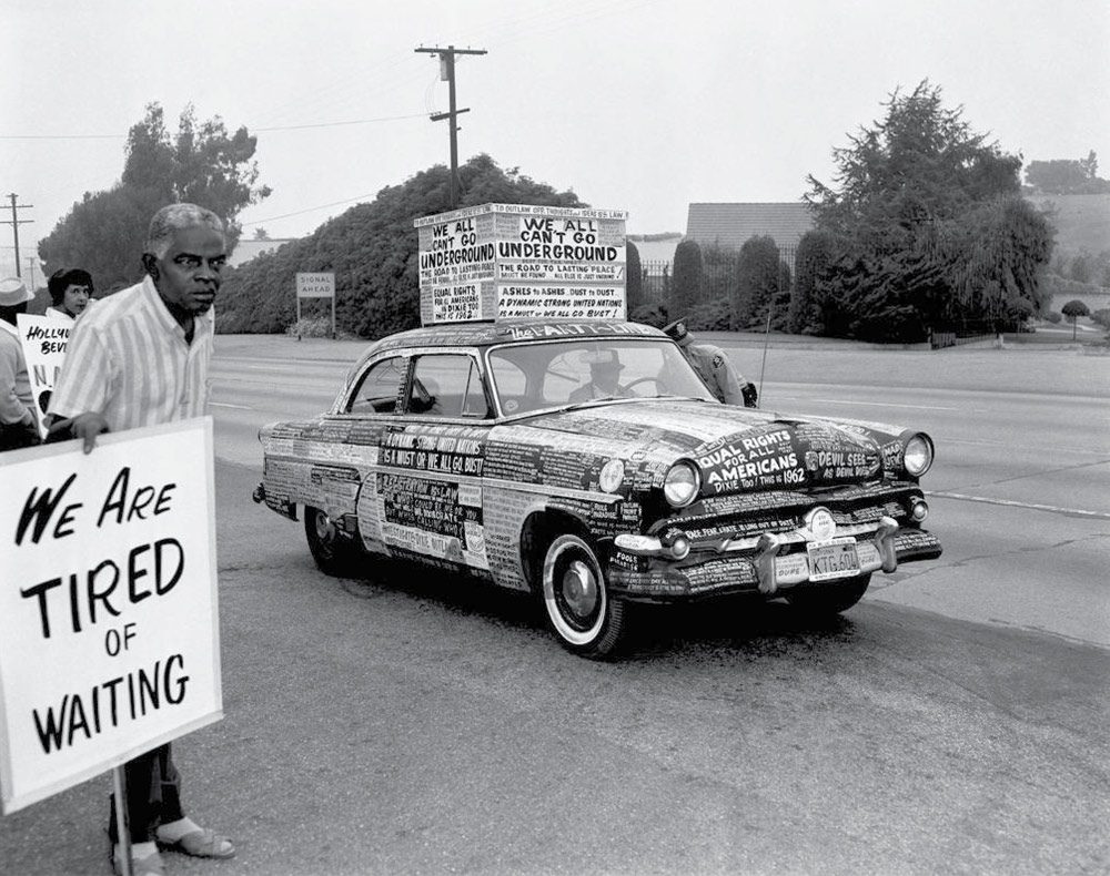 Black and white photo of a Black man holding a "WE ARE TIRED OF WAITING" sign beside a car covered in protest messages.