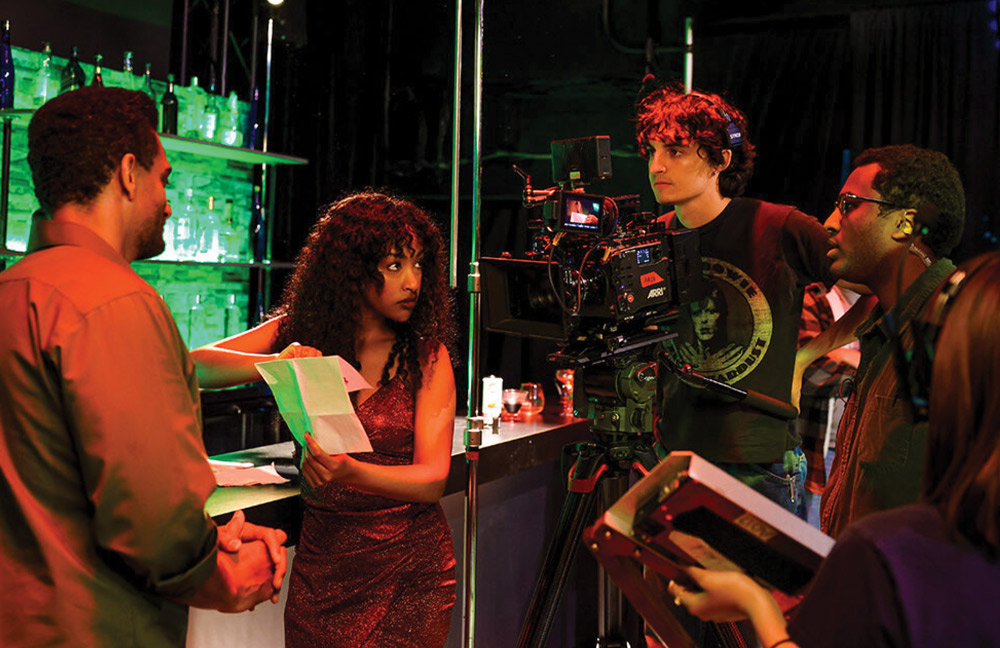 A woman in a red dress holding a script on a film set, surrounded by crew, professional lighting, and a camera operator.