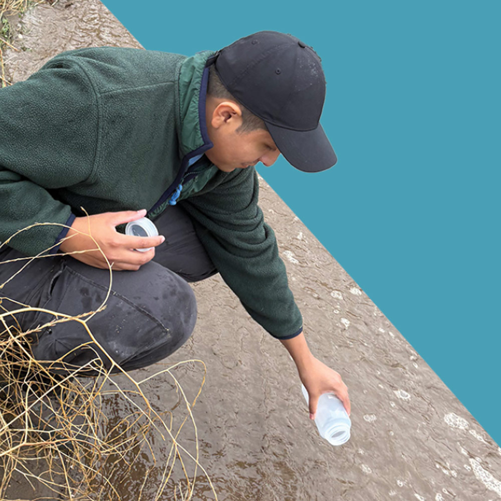A person wearing a green fleece jacket and black cap crouches by a waterway, holding a sample bottle to collect water.