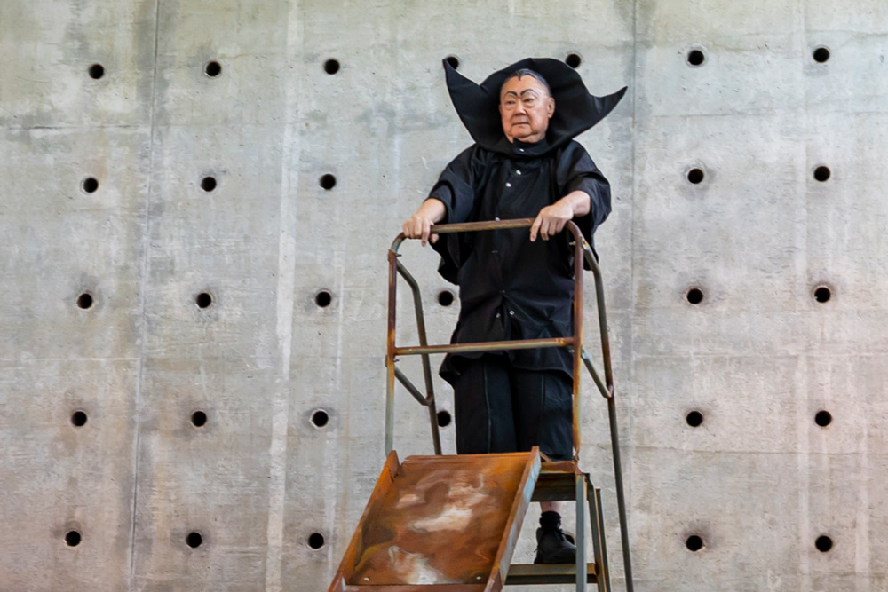 A person in a black, dramatic costume stands on a rusty metal ladder against a concrete wall with circular indentations.