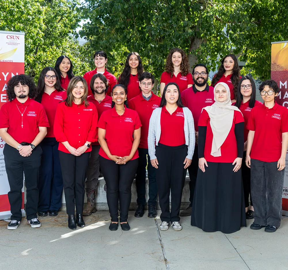 A large group of students and staff wearing matching red CSUN polo shirts posing together outdoors in front of trees.