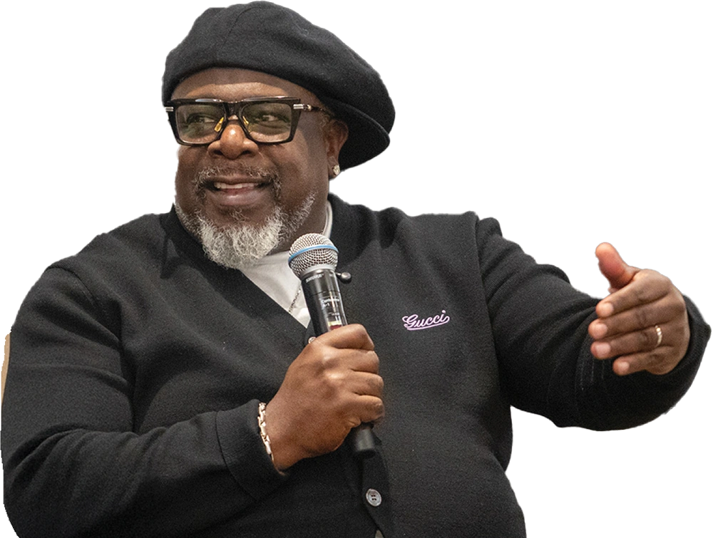 Man in a black beret and cardigan with "Gucci" text, holding a microphone and gesturing while speaking.