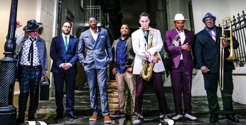Group of seven men in formal attire, including brass musicians, standing together in a dimly lit alleyway at night.